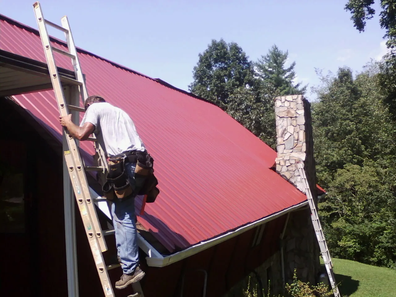 Roofer on a ladder beside a red metal roof and stone chimney — Soffit & Fascia Repair by Roofing Brothers in Grand Terrace